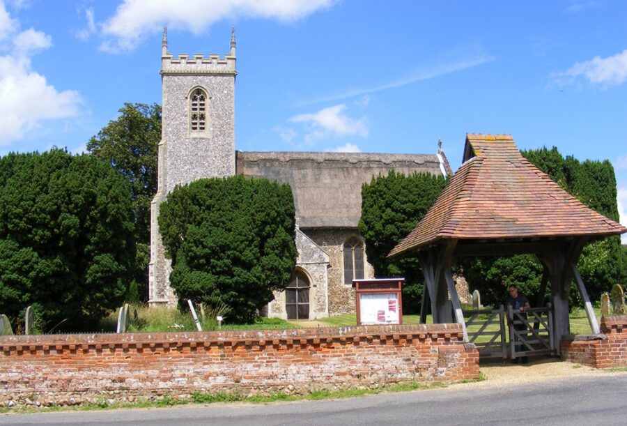 The photo shows the thatched church, lychgate and red brick wall surrounding the church on a sunny day.